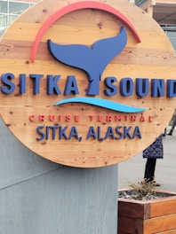 A wooden circular sign mounted on a concrete base featuring the text 'Sitka Sound Cruise Terminal Sitka, Alaska' in blue and red lettering. The sign includes a stylized whale tail graphic and a curved red accent above the text. There are partially visible people in the background along with a building.