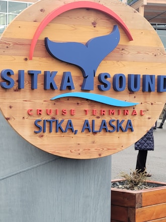 A wooden circular sign mounted on a concrete base featuring the text 'Sitka Sound Cruise Terminal Sitka, Alaska' in blue and red lettering. The sign includes a stylized whale tail graphic and a curved red accent above the text. There are partially visible people in the background along with a building.