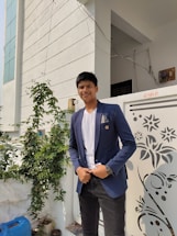 A young person stands outside near a gated entrance with decorative patterns. They are wearing a dark blue blazer, white shirt, and a wristwatch. Green plants are visible in the background, along with a partial view of a modern building with white walls and geometric lines.