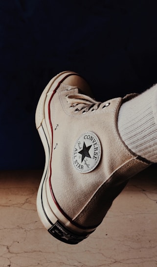 A beige Converse All Star high-top sneaker is prominently displayed against a dark background with a light-colored floor. The classic Converse logo patch is visible on the side of the shoe, alongside the shoe's white laces and a red stripe running along the sole.