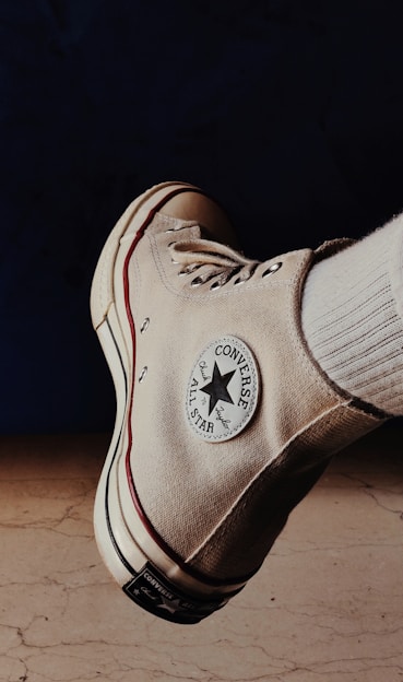 A beige Converse All Star high-top sneaker is prominently displayed against a dark background with a light-colored floor. The classic Converse logo patch is visible on the side of the shoe, alongside the shoe's white laces and a red stripe running along the sole.