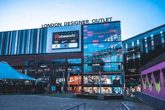 A modern building with large glass windows and multiple brand logos, including Cineworld, Nike, Adidas, and Calvin Klein, displayed on electronic screens. The architecture features sleek, vertical lines and extensive use of metallic and glass materials. The area includes outdoor seating and a partially visible tent structure.