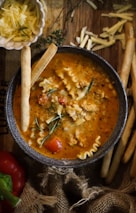 A bowl of hearty soup with a vibrant orange broth, small pasta pieces, ground meat, and fresh herbs sits on a wooden surface. Grated cheese is sprinkled on top, and breadsticks are placed to the side. Nearby, a small dish contains more shredded cheese, and a red pepper is partially visible. The setup includes rustic burlap fabric for a cozy presentation.