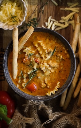 A bowl of hearty soup with a vibrant orange broth, small pasta pieces, ground meat, and fresh herbs sits on a wooden surface. Grated cheese is sprinkled on top, and breadsticks are placed to the side. Nearby, a small dish contains more shredded cheese, and a red pepper is partially visible. The setup includes rustic burlap fabric for a cozy presentation.