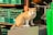 A ginger and white cat sits on a large plastic container surrounded by green plastic crates and various metal tools. The cat looks curious and attentive.