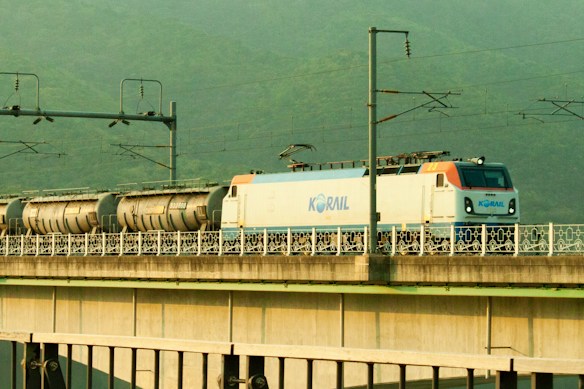 A train with the logo 'Korail' is traveling on a bridge. The train consists of a white and blue engine with several cylindrical tanker cars following it. Overhead electric cables and supporting structures are visible above the train. The background features a lush, green hilly landscape.