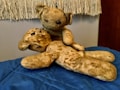 Two worn and vintage teddy bears are placed on a blue quilted surface. One bear is sitting upright, leaning over the other bear, which is lying down. The stuffed animals have a well-loved and aged appearance, with visible wear on their fabric and patches.