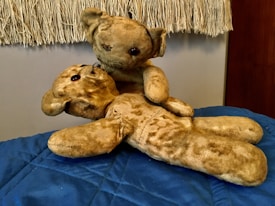 Two worn and vintage teddy bears are placed on a blue quilted surface. One bear is sitting upright, leaning over the other bear, which is lying down. The stuffed animals have a well-loved and aged appearance, with visible wear on their fabric and patches.