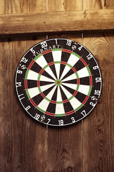 A dartboard is mounted on a wooden wall. The board has black, white, red, and green segments with numbers around the edge. The wall appears rustic, with visible wood grain and knots.