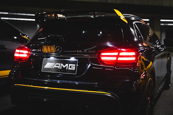 A rear view of a black Mercedes-Benz GLA 45 AMG with tail lights illuminated in a dark parking area. The vehicle features a badge and a sticker with foreign characters. The setting appears to be indoors with artificial lighting reflecting off the car's surface.