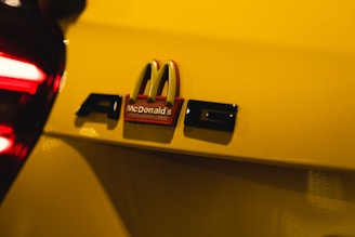 A close-up of a car with a prominent gray AMG badge beside a McDonald's logo, set against a bright yellow background. The image highlights the contrast between the brand logos.