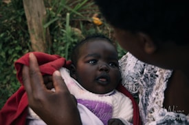 A baby wrapped in a warm red blanket gazes up at an adult, who is holding them closely. The background displays some green foliage, suggesting an outdoor setting.