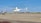 A white helicopter is hovering above a runway with a city skyline in the background. Another helicopter is parked to the left. The scene is set in a clear, blue-sky day.