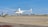 A white helicopter is hovering above a runway with a city skyline in the background. Another helicopter is parked to the left. The scene is set in a clear, blue-sky day.