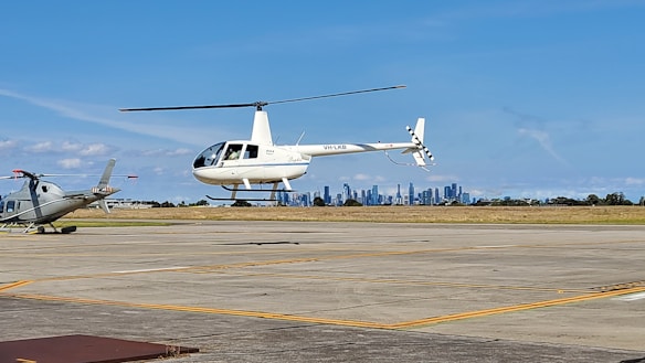 A white helicopter is hovering above a runway with a city skyline in the background. Another helicopter is parked to the left. The scene is set in a clear, blue-sky day.