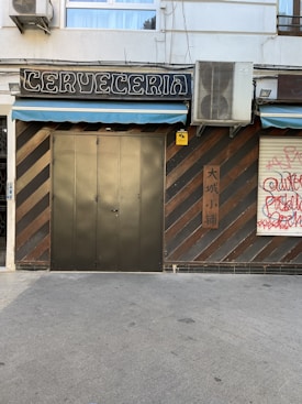 A closed establishment with a sign reading 'Cerveceria' above a metal door. The facade has a striped design with alternating brown and dark colors. An air conditioning unit is mounted above, next to the sign. There's graffiti on the adjacent shutter, and a sign with Asian characters on the wall.
