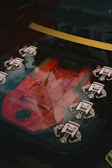 A close-up of a car engine through a transparent panel, featuring a red AMG engine with several stylized decals of cartoonish faces with flexed arms. The background shows reflections of trees, indicating the setting is outdoors during the daytime.
