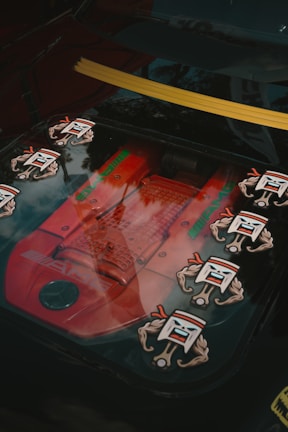 A close-up of a car engine through a transparent panel, featuring a red AMG engine with several stylized decals of cartoonish faces with flexed arms. The background shows reflections of trees, indicating the setting is outdoors during the daytime.