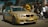 A gold-colored BMW car is prominently displayed in an outdoor setting with several people gathered around, suggesting a car event or meetup. The car has custom blue text on the side and red-blue stripes on the front. The people are casually dressed, and the scene appears lively and social.