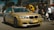 A gold-colored BMW car is prominently displayed in an outdoor setting with several people gathered around, suggesting a car event or meetup. The car has custom blue text on the side and red-blue stripes on the front. The people are casually dressed, and the scene appears lively and social.