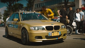 A gold-colored BMW car is prominently displayed in an outdoor setting with several people gathered around, suggesting a car event or meetup. The car has custom blue text on the side and red-blue stripes on the front. The people are casually dressed, and the scene appears lively and social.