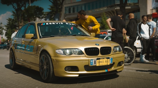 A gold-colored BMW car is prominently displayed in an outdoor setting with several people gathered around, suggesting a car event or meetup. The car has custom blue text on the side and red-blue stripes on the front. The people are casually dressed, and the scene appears lively and social.