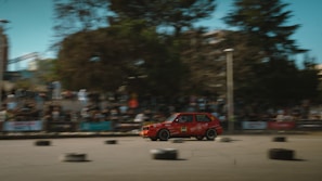 A red car speeds through a race track surrounded by an audience in bleachers. The image captures the motion of the vehicle with a blur effect. Black tires are scattered on the pavement serving as obstacles or markers. Trees and buildings in the background add a sense of an urban environment.