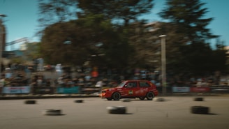 A red car speeds through a race track surrounded by an audience in bleachers. The image captures the motion of the vehicle with a blur effect. Black tires are scattered on the pavement serving as obstacles or markers. Trees and buildings in the background add a sense of an urban environment.