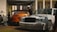 Two cars are parked under a canopy. The car on the left is an orange Toyota Supra with a large wing, while the car on the right is a white Mercedes with decals. Several people are nearby, and the setting appears to be an outdoor car event in an urban area with modern buildings in the background.