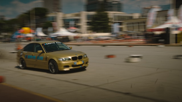 A gold BMW car is captured in mid-drift on an urban road, with blurred motion effects highlighting high speed. It is surrounded by a makeshift track marked by small barrels. Buildings and event tents are visible in the background, suggesting a motorsport event.