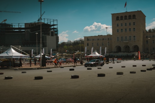 A scene depicting an outdoor event with several tents and people milling about, possibly for a car show or race, as there are numerous tires arranged on the ground. A prominent beige building with arched windows is visible in the background, and another partially constructed building is seen on the left. The sky is clear with scattered clouds.