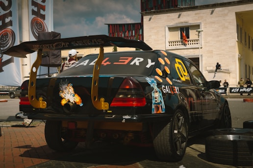A customized black car with a large rear spoiler, anime-style decals, and decorative stickers, parked on a street with various promotional banners and buildings in the background.