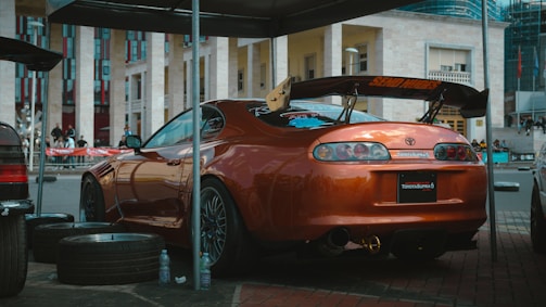 A parked sports car with a glossy orange finish is shown, featuring a prominent rear spoiler and customized decals. It is surrounded by car tires and plastic bottles, under a makeshift shelter. The setting appears to be an outdoor event in an urban area with a modern building in the background.