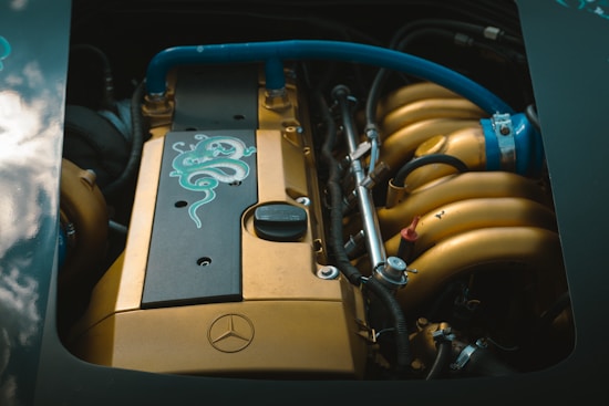 A detailed view of a car engine featuring gold and black components with blue accents. The engine has a green serpent-like design on a black cover and a Mercedes logo is visible. Various pipes, wires, and metal parts are surrounding the engine.