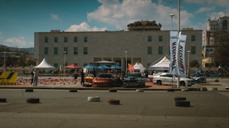 A car show or event is set up in an urban area, featuring several parked cars with modified features and accessories. There are white and red tents and banners, suggesting different brands or sponsors. The setting includes a modern building in the background with a blue sky and scattered clouds overhead. Attendees are visible walking and observing the cars.