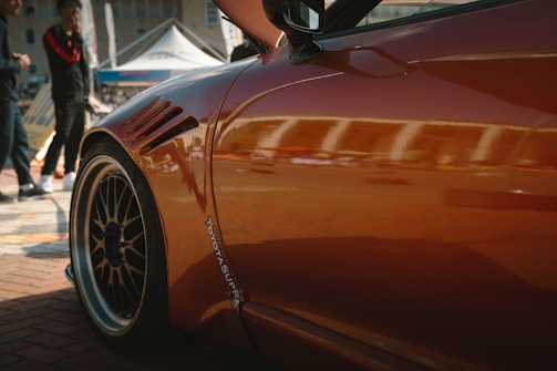 A close-up view of the side of an orange sports car, with reflections visible on its glossy surface. The car is parked on a brick road at an outdoor event, with several people standing nearby under a tent. The wheel is prominently featured in the foreground, highlighting intricate rim details.