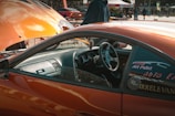 An orange car with an open hood is parked in a busy outdoor setting. The driver's side door is open, revealing a sleek, modified interior. A person stands near the hood, wearing a dark jacket. Various stickers and decals are visible on the car's windows.