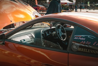 An orange car with an open hood is parked in a busy outdoor setting. The driver's side door is open, revealing a sleek, modified interior. A person stands near the hood, wearing a dark jacket. Various stickers and decals are visible on the car's windows.
