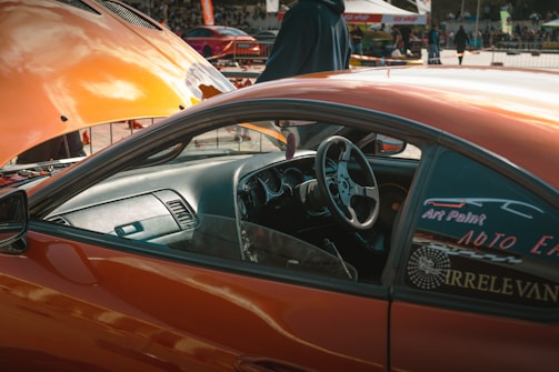 An orange car with an open hood is parked in a busy outdoor setting. The driver's side door is open, revealing a sleek, modified interior. A person stands near the hood, wearing a dark jacket. Various stickers and decals are visible on the car's windows.