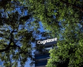 A building with the word 'Cognizant' prominently displayed on it is framed by green tree leaves. The sky is a clear blue, creating a contrast against the foliage and the building.