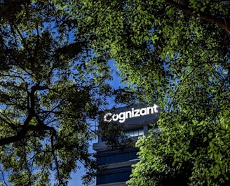 A building with the word 'Cognizant' prominently displayed on it is framed by green tree leaves. The sky is a clear blue, creating a contrast against the foliage and the building.