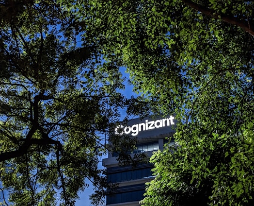 A building with the word 'Cognizant' prominently displayed on it is framed by green tree leaves. The sky is a clear blue, creating a contrast against the foliage and the building.