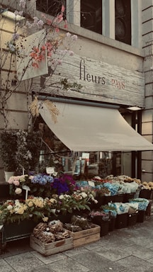 A charming flower shop with a display of vibrant, colorful flowers in various pots and containers. The shop's sign reads 'fleurs de PAYS' and is adorned with decorative branches and leaves. The awning is partially extended over the entrance.