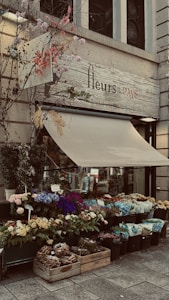 A charming flower shop with a display of vibrant, colorful flowers in various pots and containers. The shop's sign reads 'fleurs de PAYS' and is adorned with decorative branches and leaves. The awning is partially extended over the entrance.