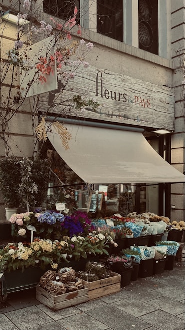 A charming flower shop with a display of vibrant, colorful flowers in various pots and containers. The shop's sign reads 'fleurs de PAYS' and is adorned with decorative branches and leaves. The awning is partially extended over the entrance.