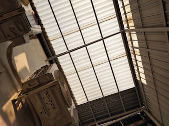 A metal corrugated roof structure with sunlight filtering through. There are two large air conditioning units mounted below the roof, with visible ductwork and vents. The shadow of the roof panels creates a pattern on the floor.