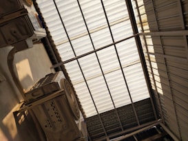 A metal corrugated roof structure with sunlight filtering through. There are two large air conditioning units mounted below the roof, with visible ductwork and vents. The shadow of the roof panels creates a pattern on the floor.
