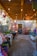 A rustic outdoor space adorned with hanging lights, colorful decorative fabrics and pots. Wooden beams support the ceiling, and a variety of vibrant crafts and plants are displayed throughout. The setting feels cozy and inviting, with a mix of natural elements and artisanal goods.
