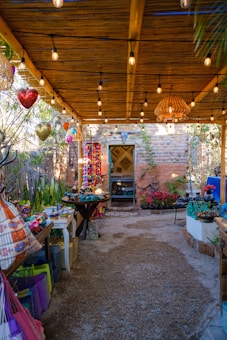 A rustic outdoor space adorned with hanging lights, colorful decorative fabrics and pots. Wooden beams support the ceiling, and a variety of vibrant crafts and plants are displayed throughout. The setting feels cozy and inviting, with a mix of natural elements and artisanal goods.