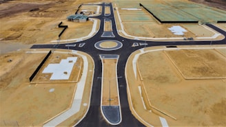 Aerial view of a newly constructed road with a roundabout in a development area. The site features patches of bare soil, concrete foundations, and partially constructed buildings. The surrounding land appears undeveloped, with empty plots of dirt and some green-covered sections indicating future construction.